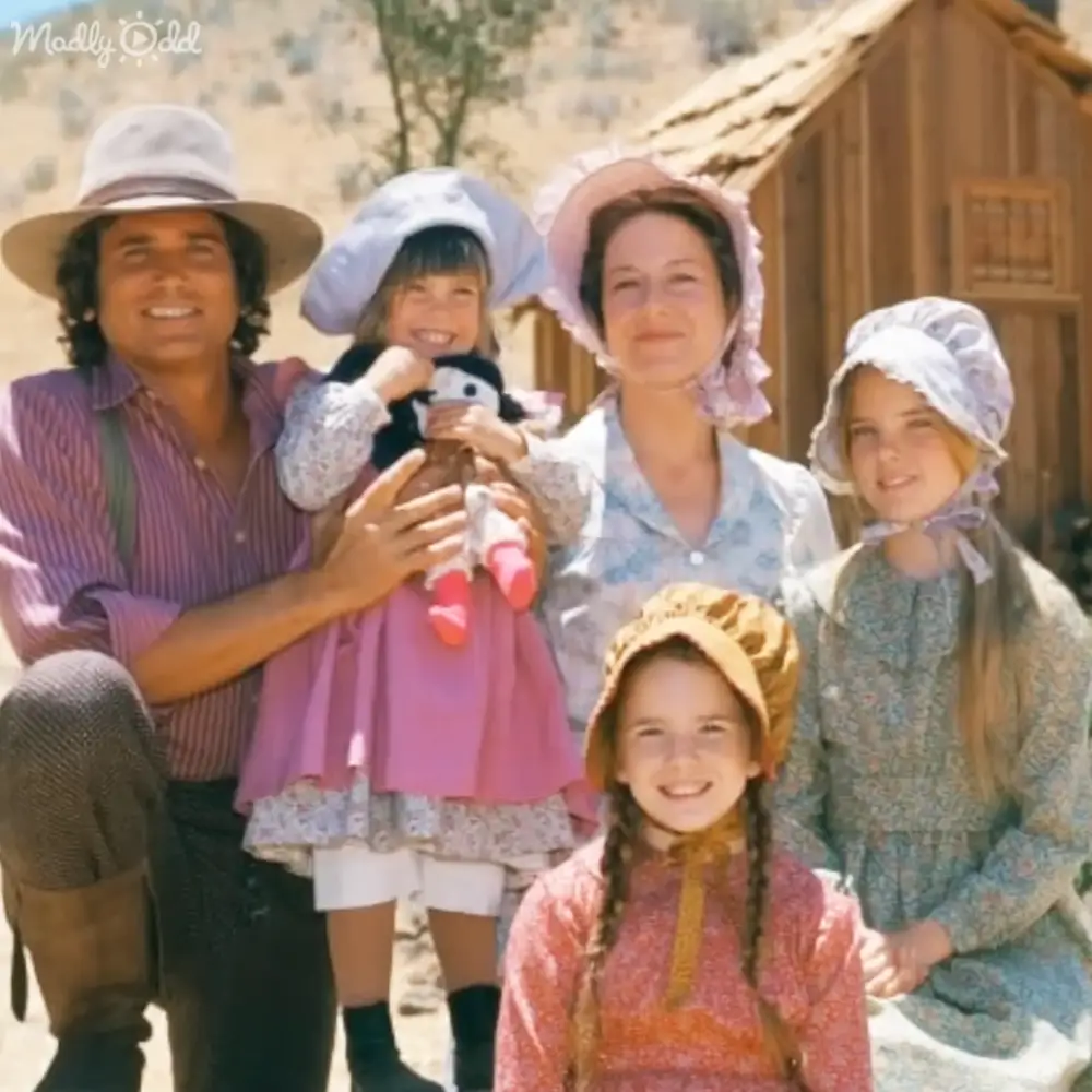 A family scene in a rustic outdoor setting with an adult male with curly hair wearing a wide-brimmed hat and striped shirt, children dressed in light-colored clothing holding dolls, and a wooden structure in the background.