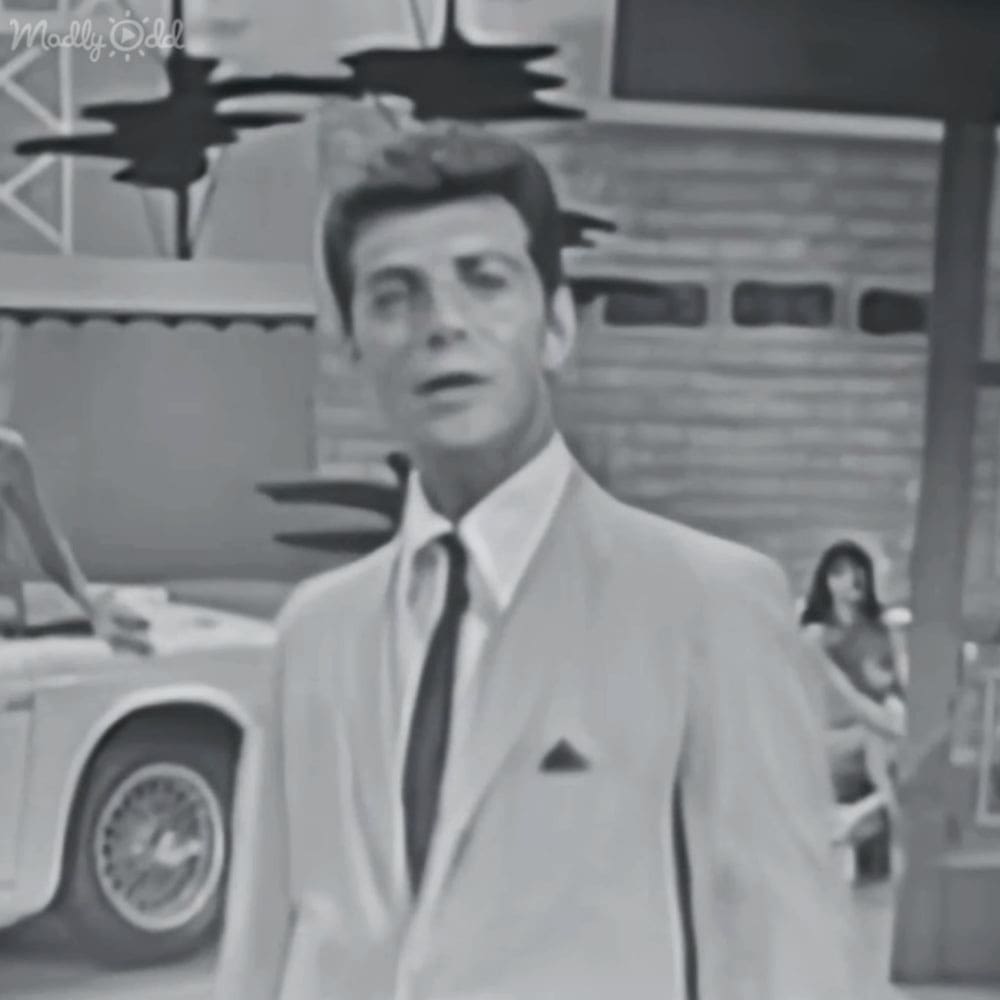 An adult male in a light-colored suit jacket stands outdoors near a vintage car with outdoor furniture and a garage door visible in the background.