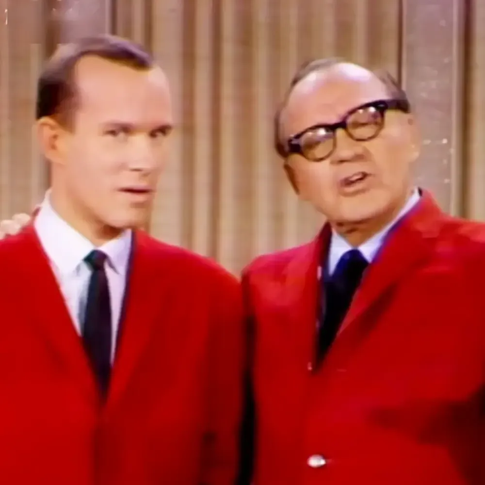 An adult male singer in a mid-20th century formal outfit stands beside an older adult male wearing a bright red blazer, both set against vertical wooden paneling typical of a studio backdrop.