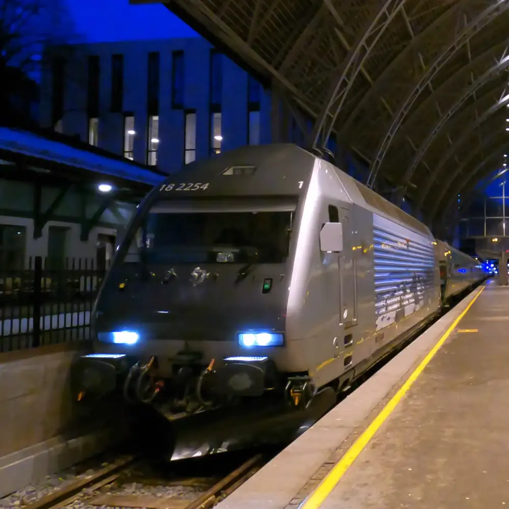 A modern electric locomotive numbered '18 2254' at a train station with arched ceilings, large windows, and yellow tactile paving along the platform in an evening or nighttime setting.