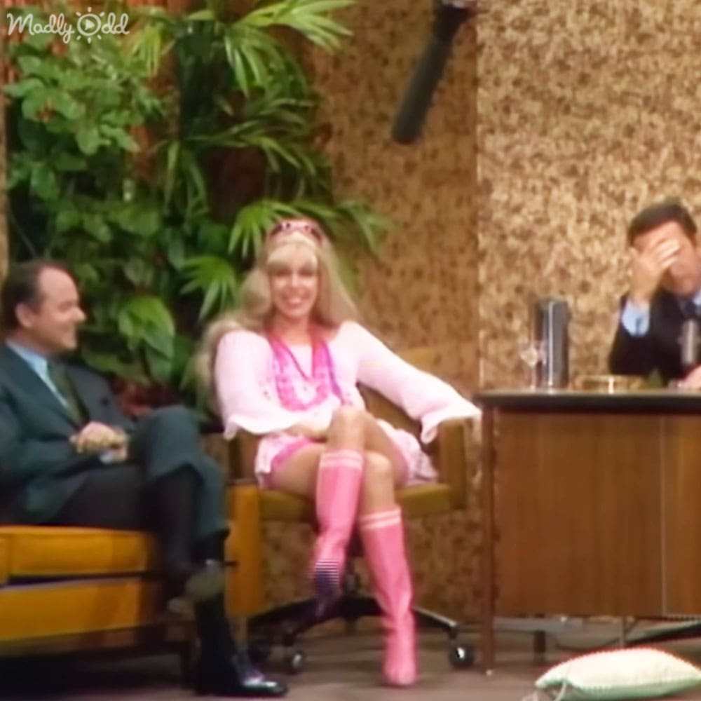 A middle-aged man in a dark suit sits on the left, while a young adult or middle-aged woman with long blonde hair and pink outfit is seated center stage. She wears a headband and necklace, suggesting she might be a guest or host of a talk show set.