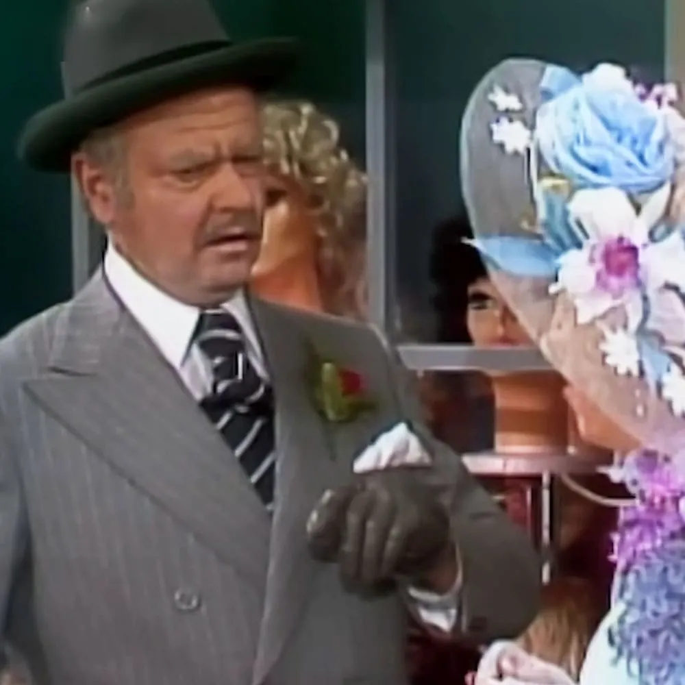 An older adult male in a formal setting with a gray pinstripe suit and black gloves, sitting next to a middle-aged female wearing a purple floral dress and an elaborate blue and white hat adorned with flowers.
