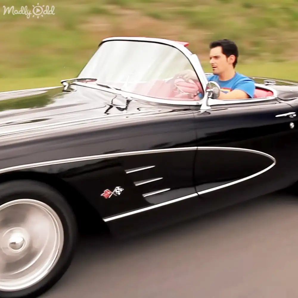 A young adult male drives a classic black convertible with chrome details, wearing a blue t-shirt and driving on an outdoor rural road.