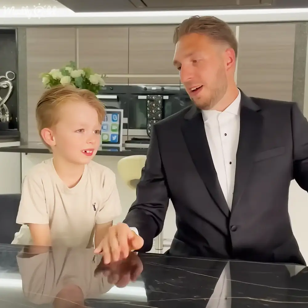 A young child and an adult male interact in a modern kitchen setting with contemporary decor, bright lighting, and greenery.