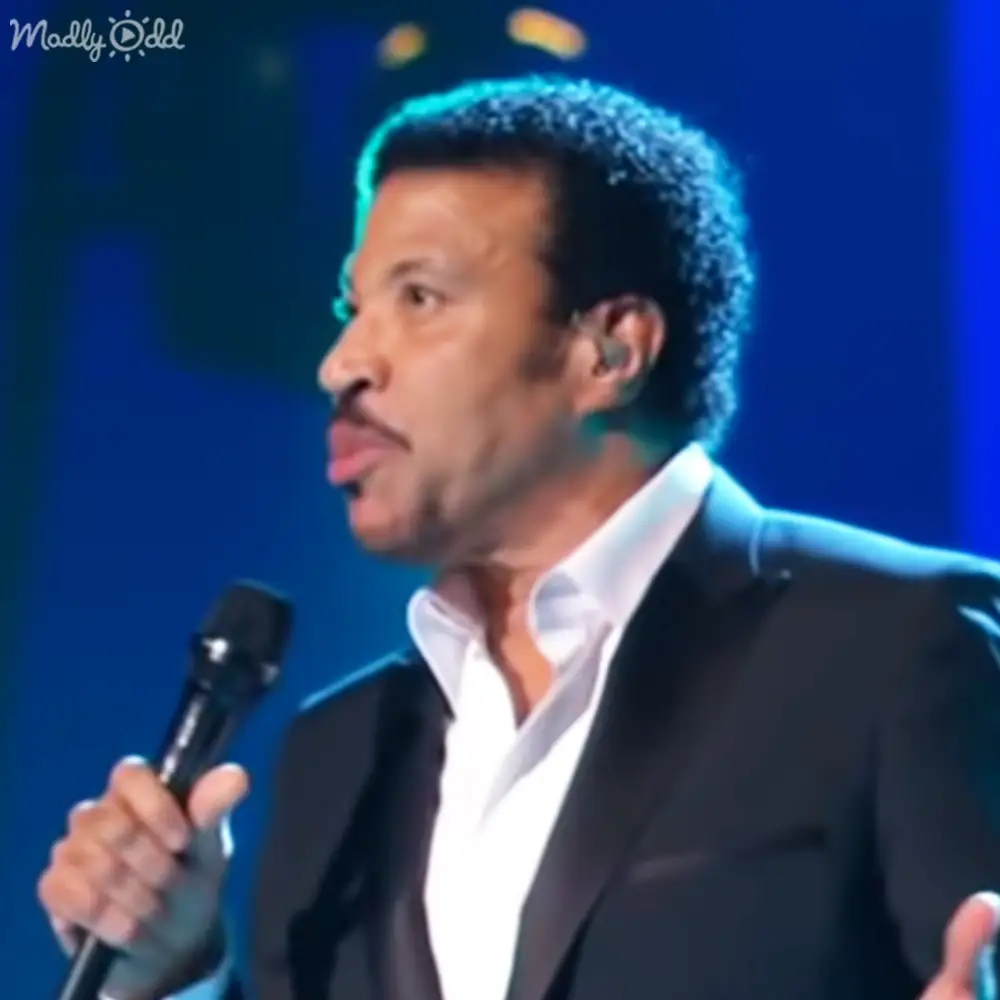 An adult male performer on stage, dressed in a black suit with white shirt and holding a microphone under blue lighting.
