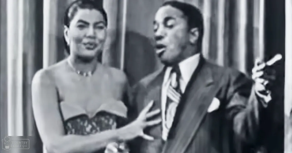 A middle-aged man in a dark suit with striped tie holds a microphone while an adult woman in a patterned dress with a low-cut neckline and beaded necklace performs indoors, likely on stage.