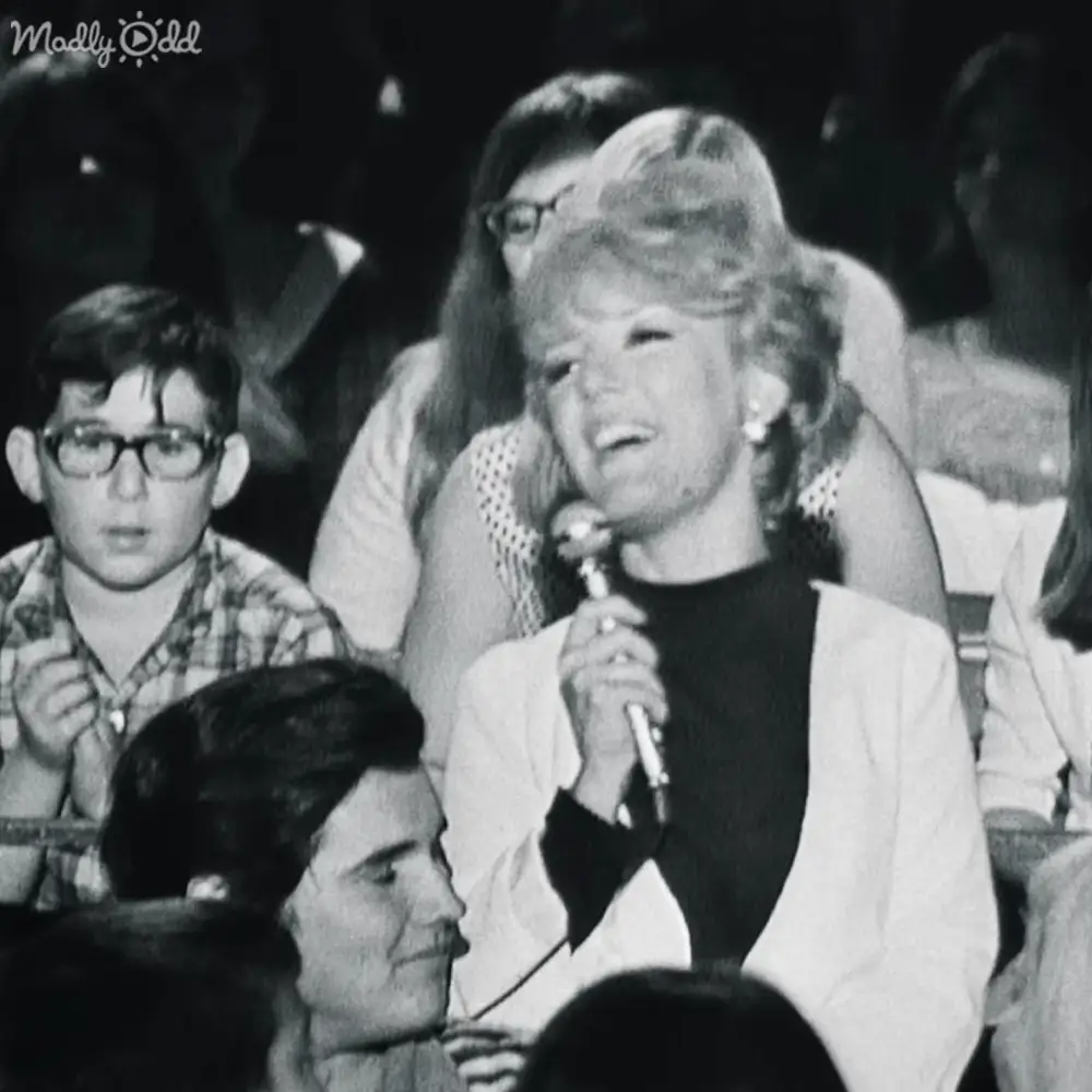 A woman singing into a microphone in what appears to be a performance hall, surrounded by seated spectators under stage lighting.
