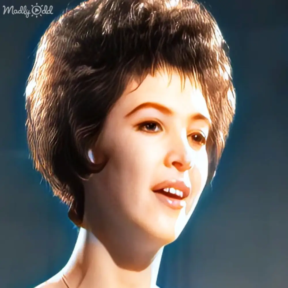 A young adult female wearing a mid-20th-century white cotton or linen garment indoors with stage lighting.