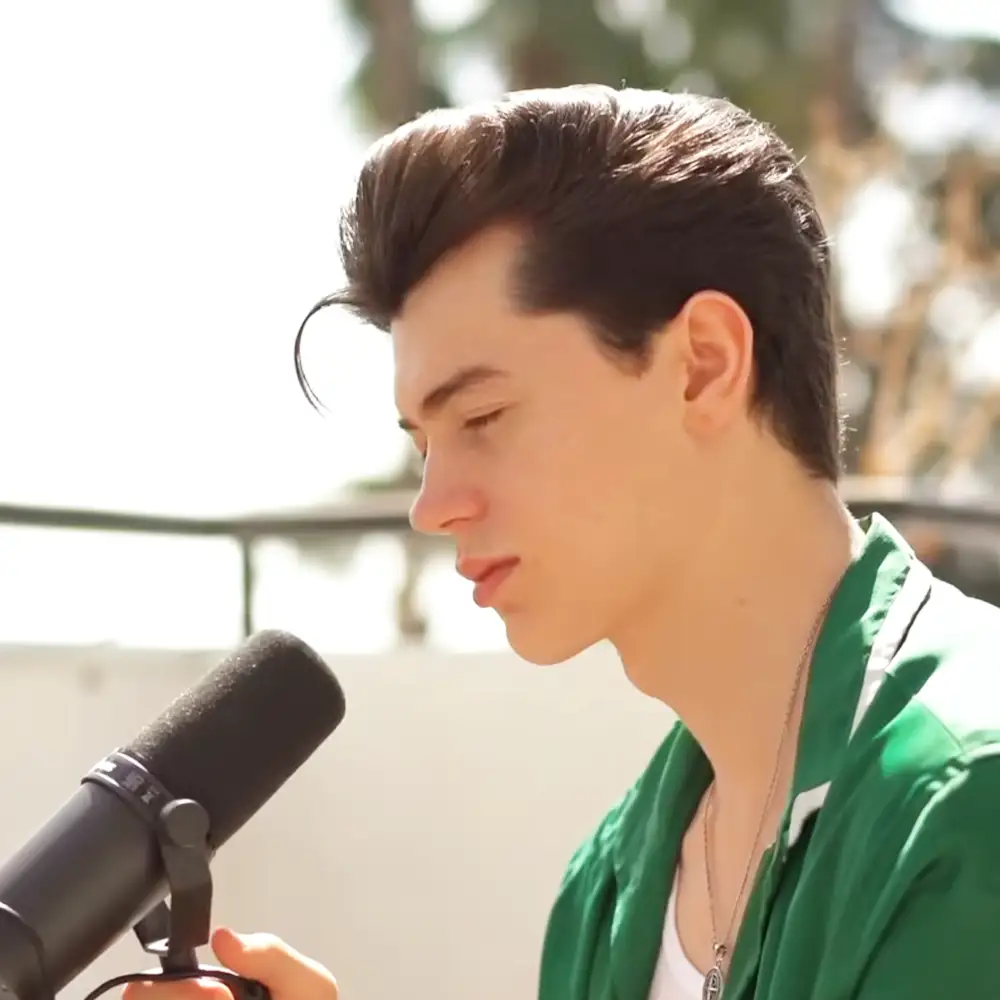 A young adult male wearing a green jacket with white inner lining, holding a microphone outdoors on a sunny day.