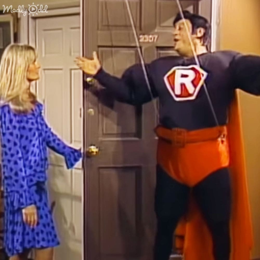 A man in a superhero costume stands beside a child wearing a blue dress with animal spots, indoors at home. A wooden door with a gold doorknob and number plate is prominently featured.