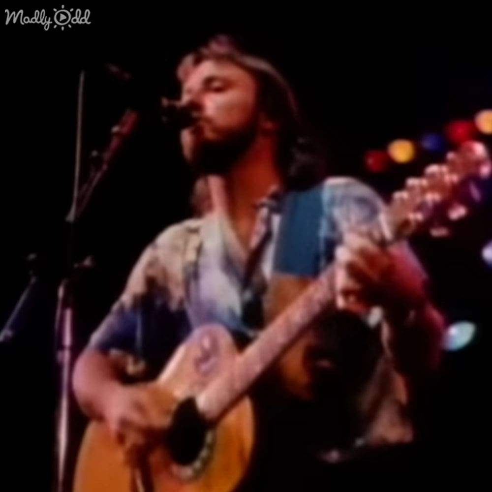 A young adult male musician wearing a loose, short-sleeved 1970s-style patterned shirt performs on stage at an indoor concert venue with blurred background lights and stage lighting.
