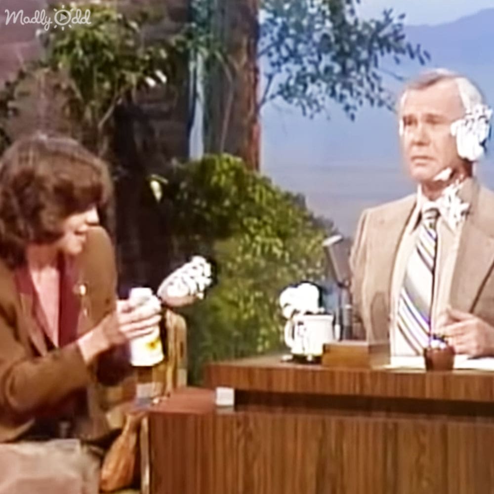 An adult female and older male appear outdoors on a talk show set, surrounded by trees and mountains; they wear formal attire with intricate patterns and accessories.
