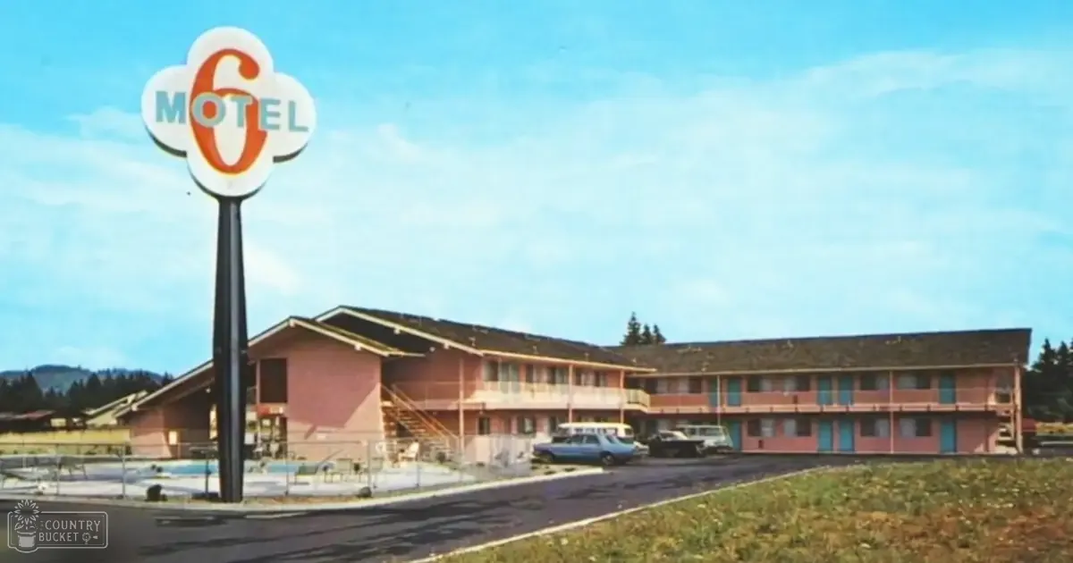 A two-story pink motel with balconies, situated in a rural area under clear skies. Trees and hills are visible in the background.