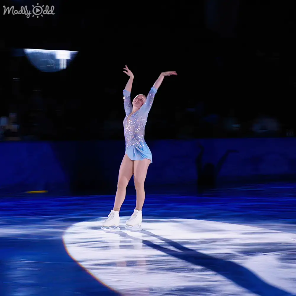 A young adult female figure skater performs on an indoor ice rink under artificial stage lighting, wearing a modern light blue and possibly silver glittery figure skating dress with long sleeves and white ice skates.