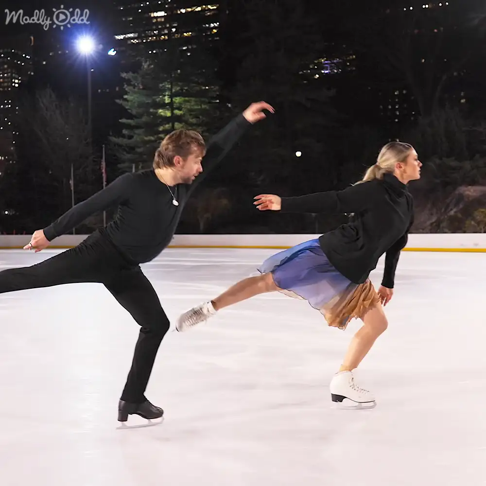 An adult male ice skater wearing a black turtleneck sweater and pants, skating at an outdoor rink with city lights and trees in the background during winter.