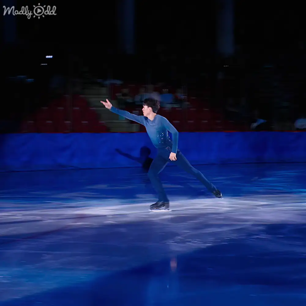A young adult male figure skater performing on an indoor ice rink with blue lighting, wearing a teal or dark blue, form-fitting garment and ice skates.