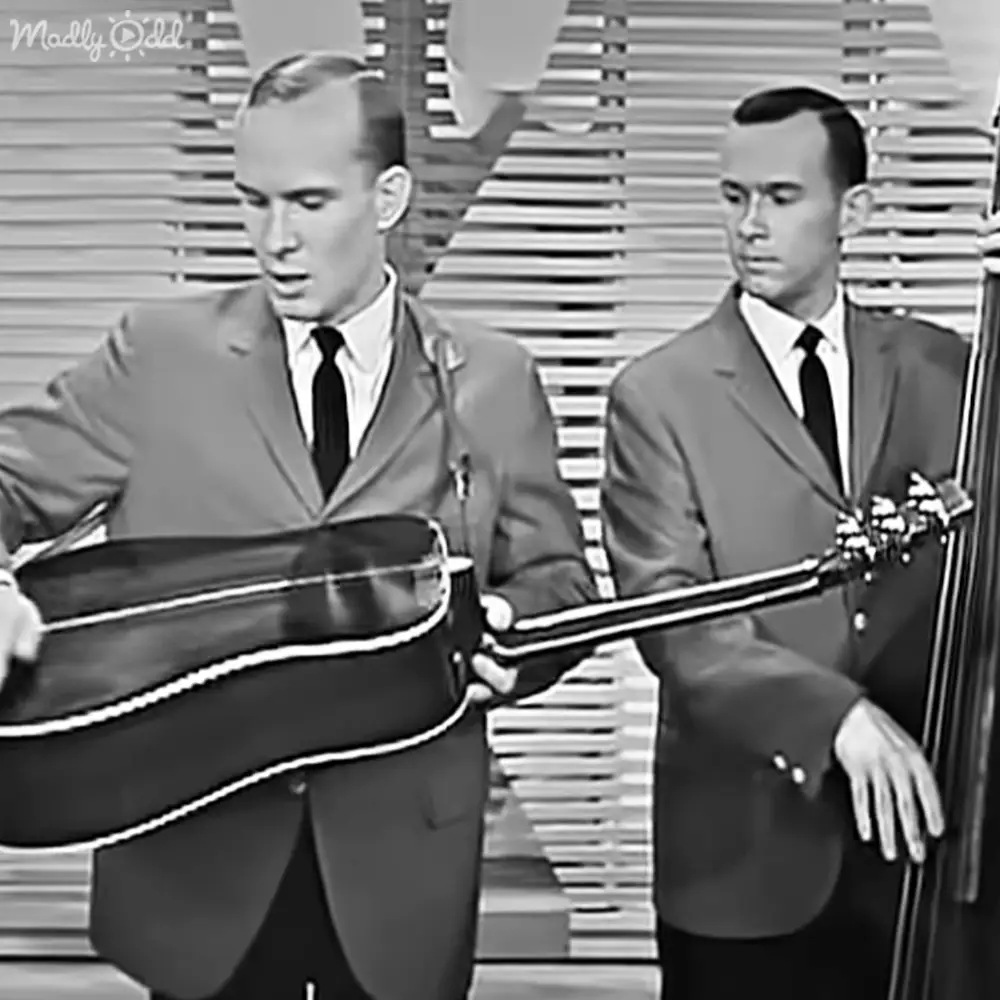 Young adult males in formal dark suits with a large stringed instrument and microphone stand on stage indoors, daytime lighting.