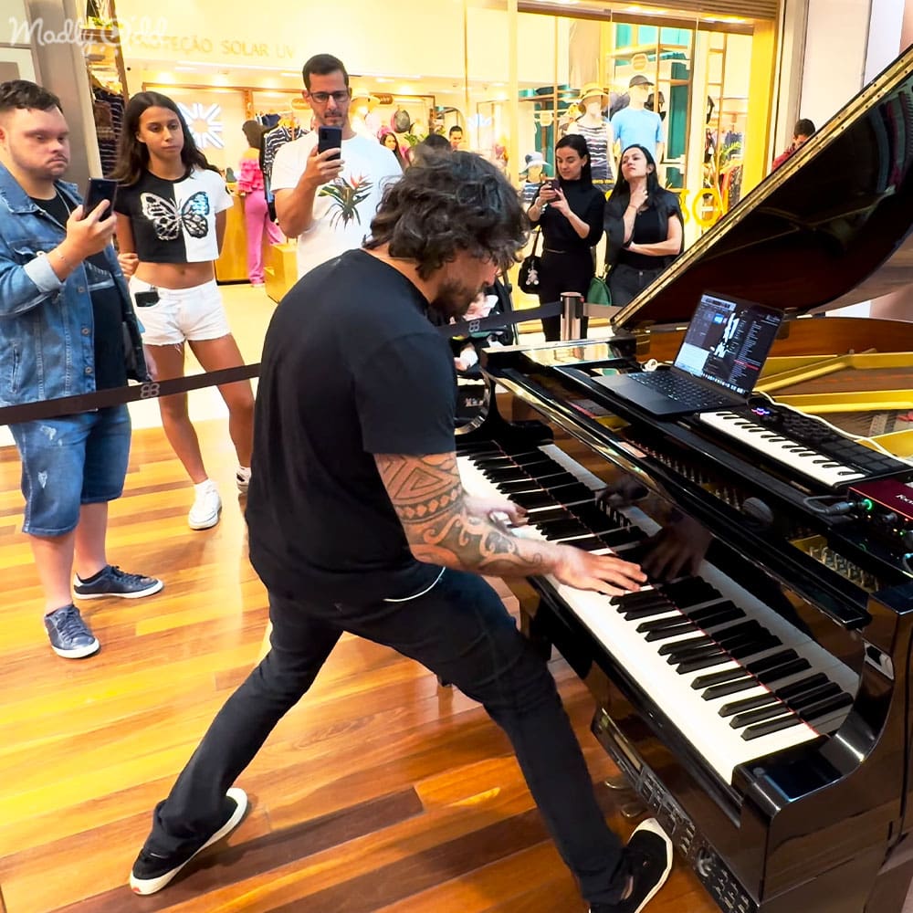 An adult male singer playing a grand piano in a shopping mall, surrounded by shoppers taking photos. Sign reads 'PROTEÇÃO SOLAR UV'.