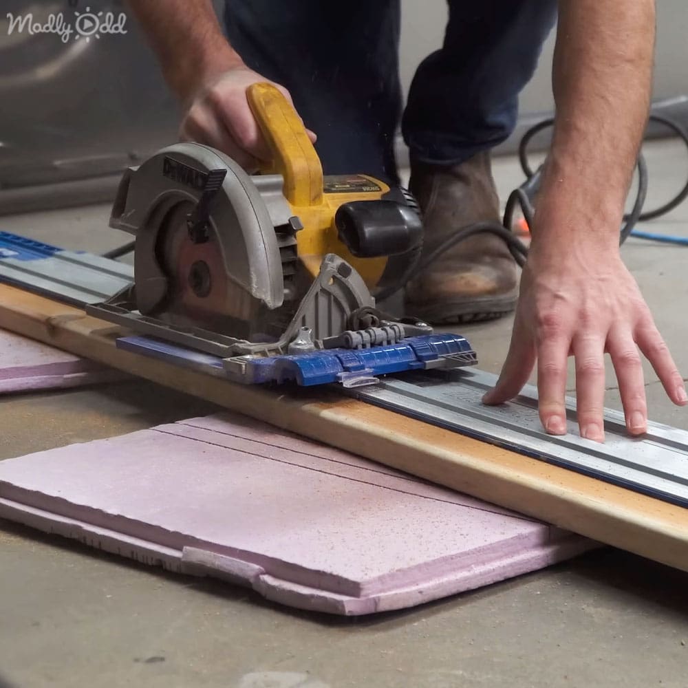 An adult male wearing dark blue jeans and brown work boots using a DeWalt brand circular saw to cut light purple foam board in an indoor workshop setting with concrete floor and coiled cables.