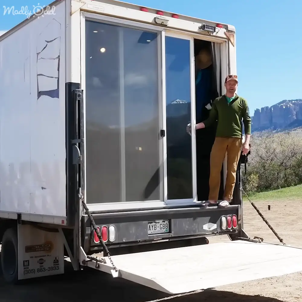 Adult male wearing a green long-sleeve polo shirt, khaki pants, and brown shoes with sunglasses on his head stands in front of a white trailer in an outdoor rural setting with mountains and trees in the background. The season is likely spring or summer.