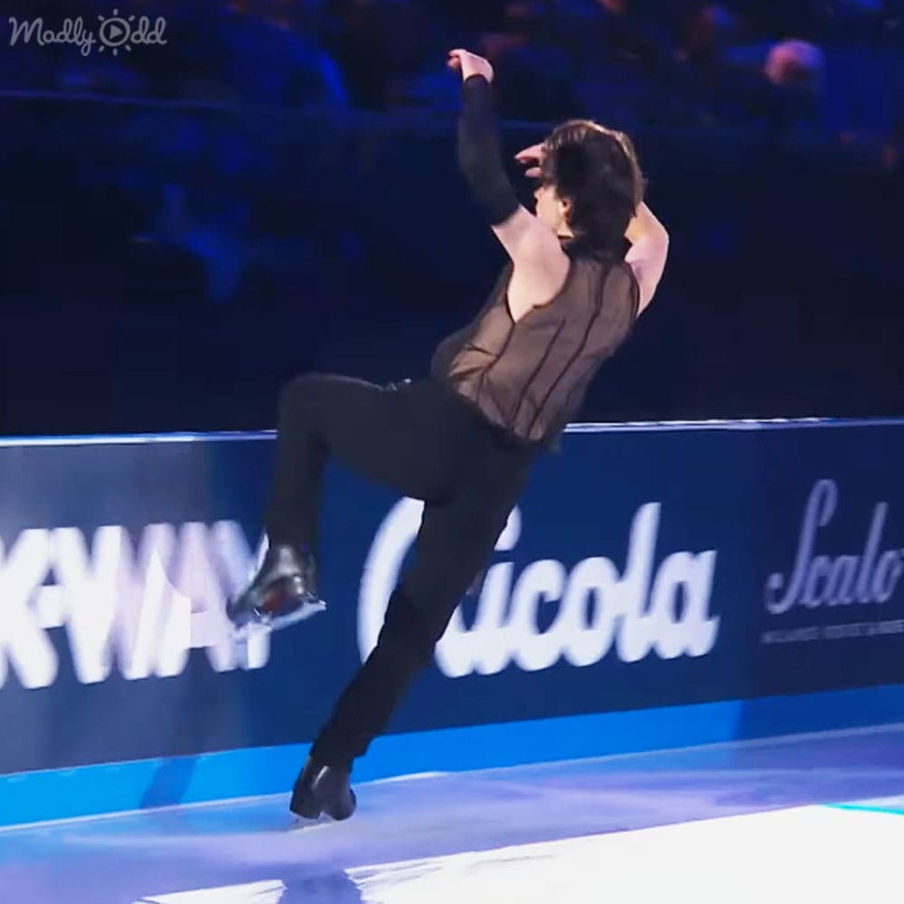 A young male figure skater performing at an indoor ice rink under artificial evening lights, with advertisements on the boards and spectators visible in the background.