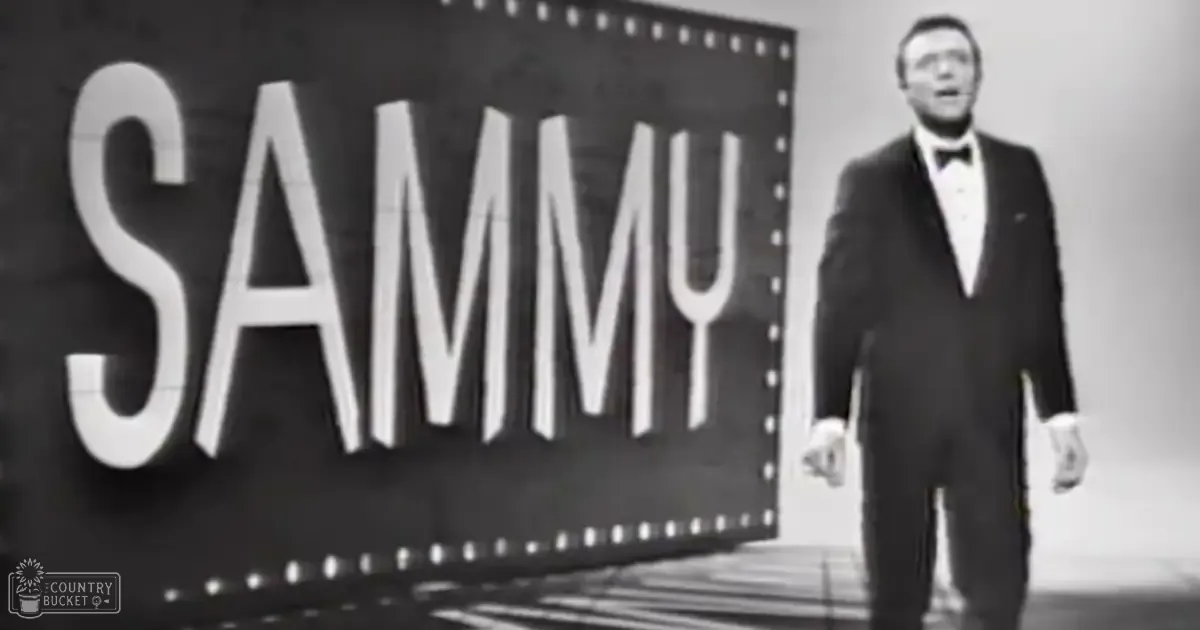 An adult male performer wearing a formal 1950s-style tuxedo in front of a 'SAMMY' sign indoors under professional stage lighting, suggesting a mid-20th century performance setting.