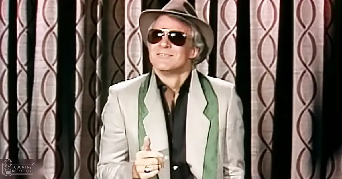 An older adult male in a formal setting, wearing a white blazer with green trim and lapels from the mid-20th century, dark sunglasses, and a light-colored fedora hat. The background features patterned curtains suggesting an indoor stage or performance area.