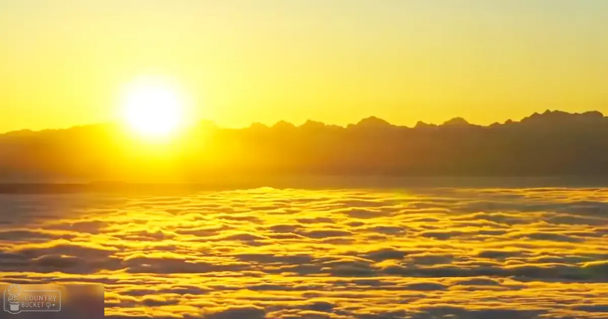 A breathtaking sunrise over rugged mountains with fluffy white clouds in a high-altitude location, bathed in golden hues and dark brown tones from early morning or late evening light.