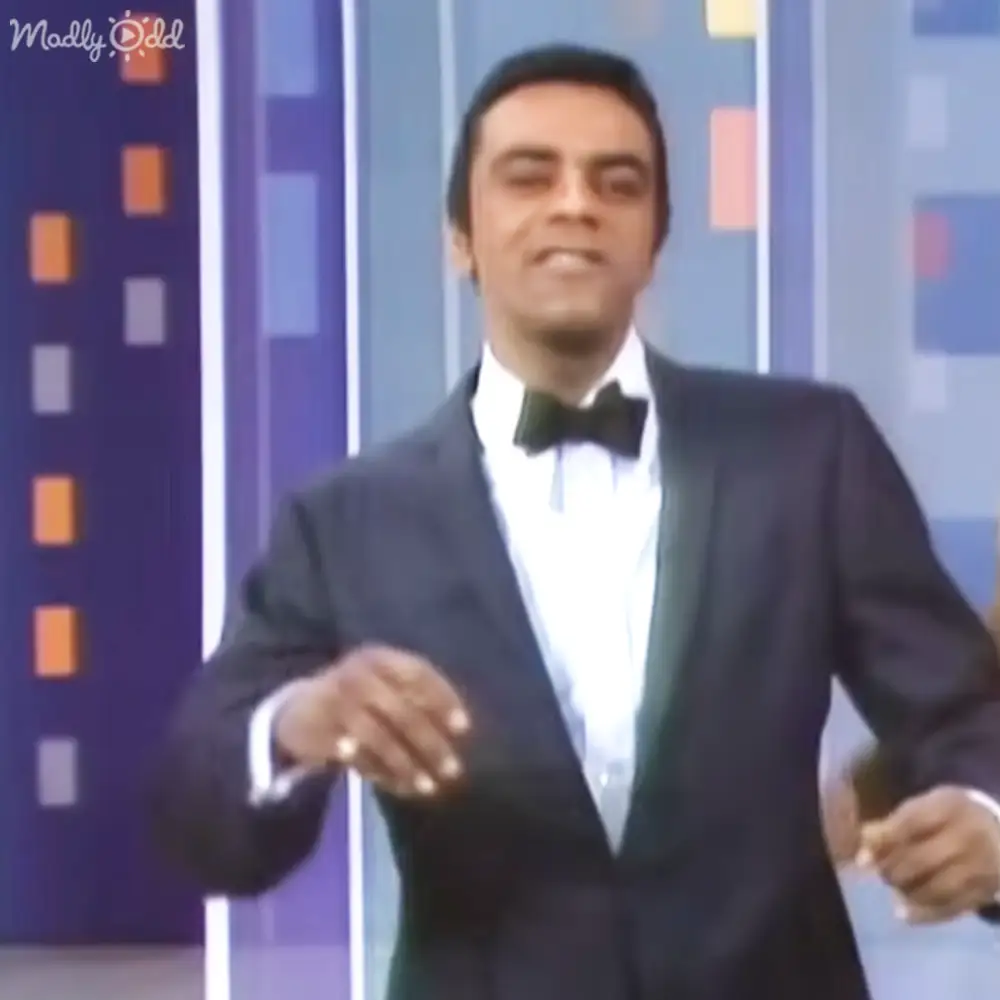 A middle-aged man wearing a formal suit with a bow tie stands indoors against a backdrop with vertical stripes of varying colors in an apparent studio setting.