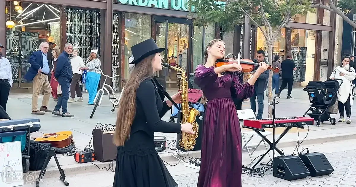 Young female musicians perform in a lively urban shopping area with red keyboard, amplifiers, and guitar case on the ground.