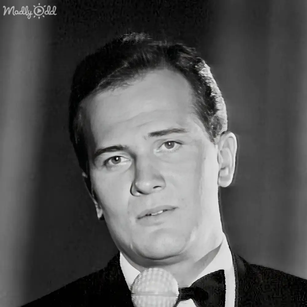 Adult male in a dark suit jacket, white shirt with collar, and bow tie; vintage style microphone and backdrop suggest mid-20th century setting.