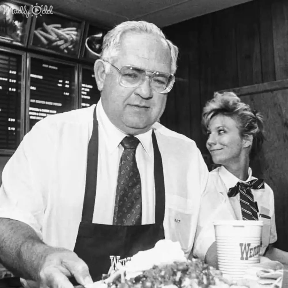 An older adult male wearing a white shirt with dark suspenders and a patterned tie stands at the counter in a fast-food setting, with menu boards and food items visible behind him.