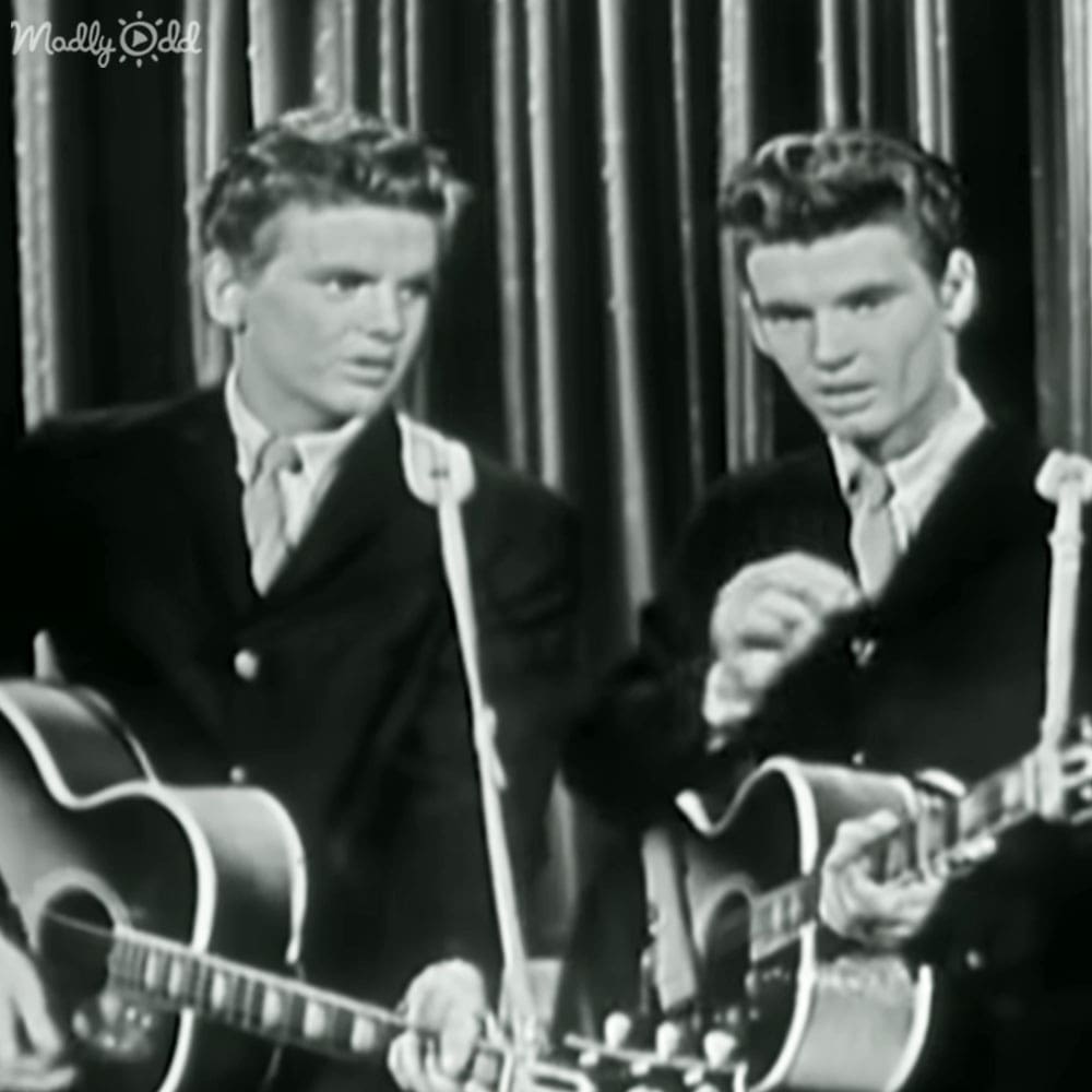 Two young adult males performing music in a studio setting with microphones and guitars.