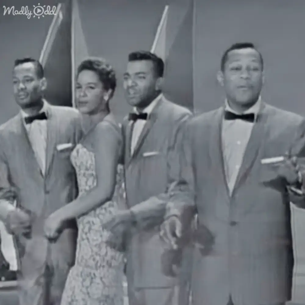 A group of performers in a mid-20th century studio setting, wearing formal suits and patterned dresses.