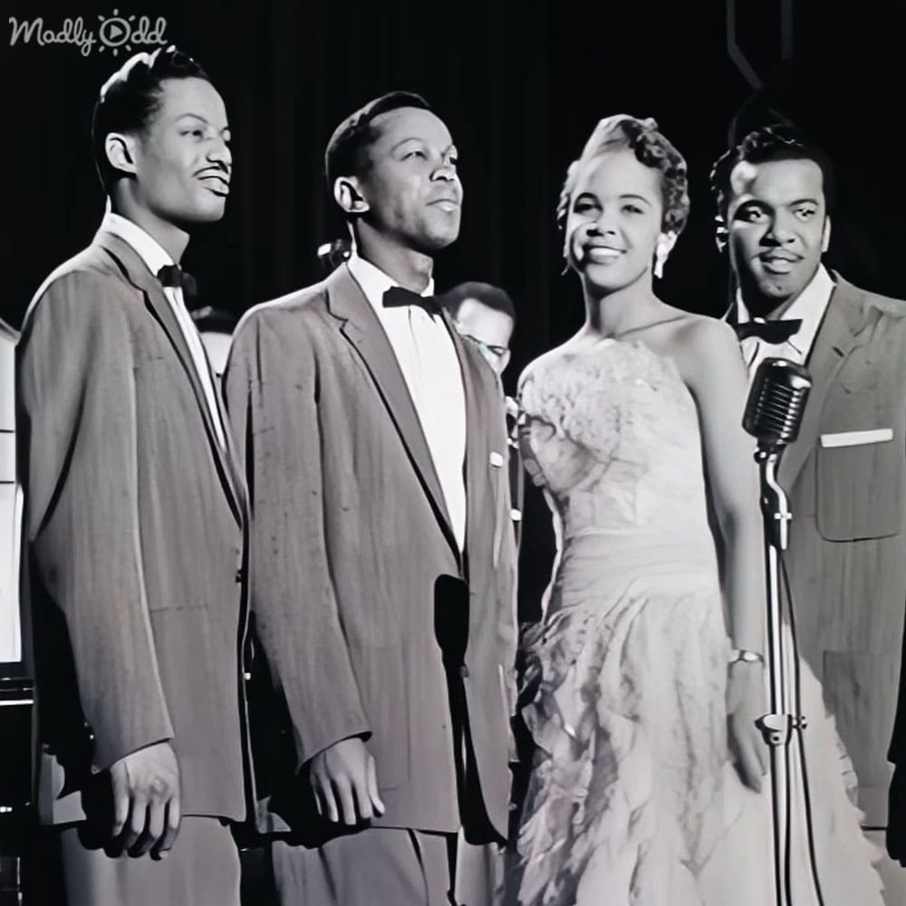 A mid-20th century musical performance scene with four individuals dressed formally in light-colored suits and a woman in a long dress, set against dark curtains or panels typical of studio or stage settings.