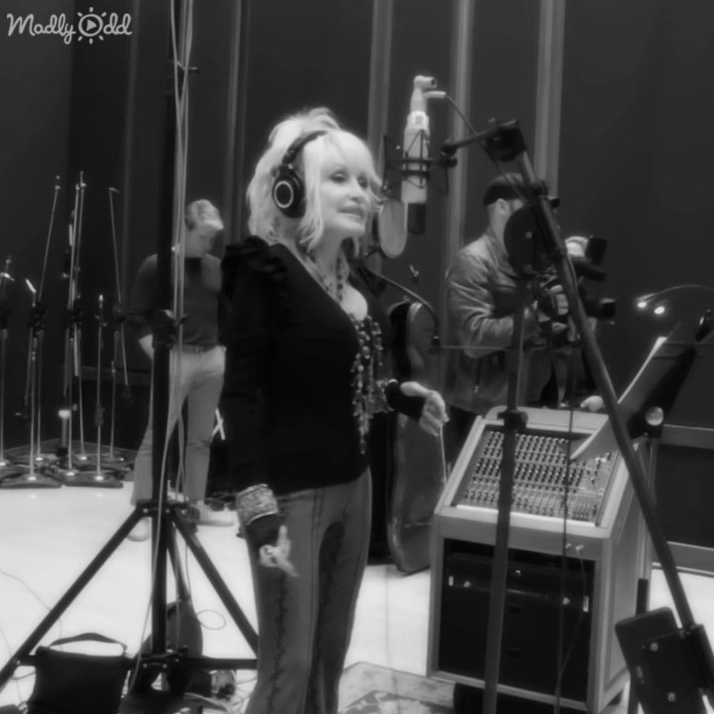 A middle-aged female in a professional recording studio, wearing a black ruffled-sleeve top with a necklace and high-waisted jeans with decorative stitching; surrounded by microphones, a soundboard, and other recording equipment.
