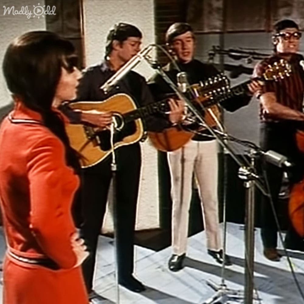 Young adult female singer in a studio, surrounded by musicians playing instruments with microphones and equipment visible in the background.