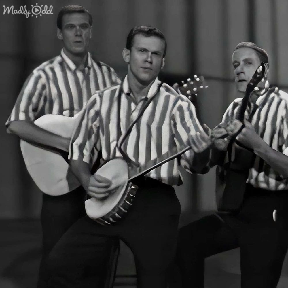 Musical performance scene with three adult males holding banjos against a stage or studio backdrop under artificial light.