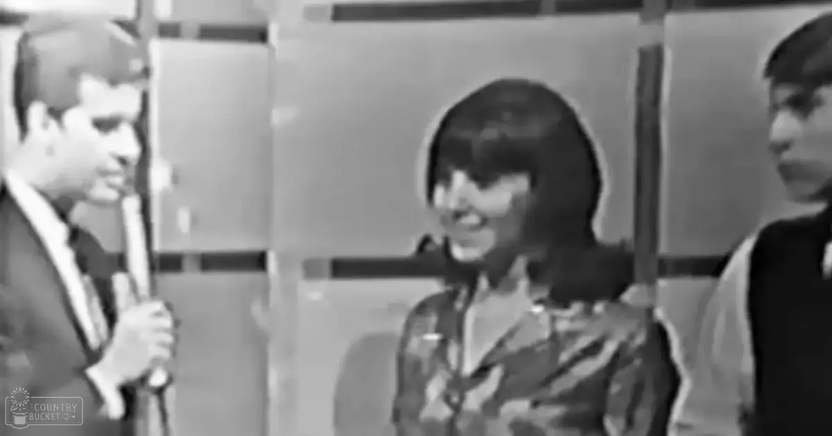 An adult male singer in a suit and bow tie holds a microphone stand; an adult female wears a patterned blouse with long sleeves; another adult male in similar attire stands beside her, all set against simple vertical paneling indoors.