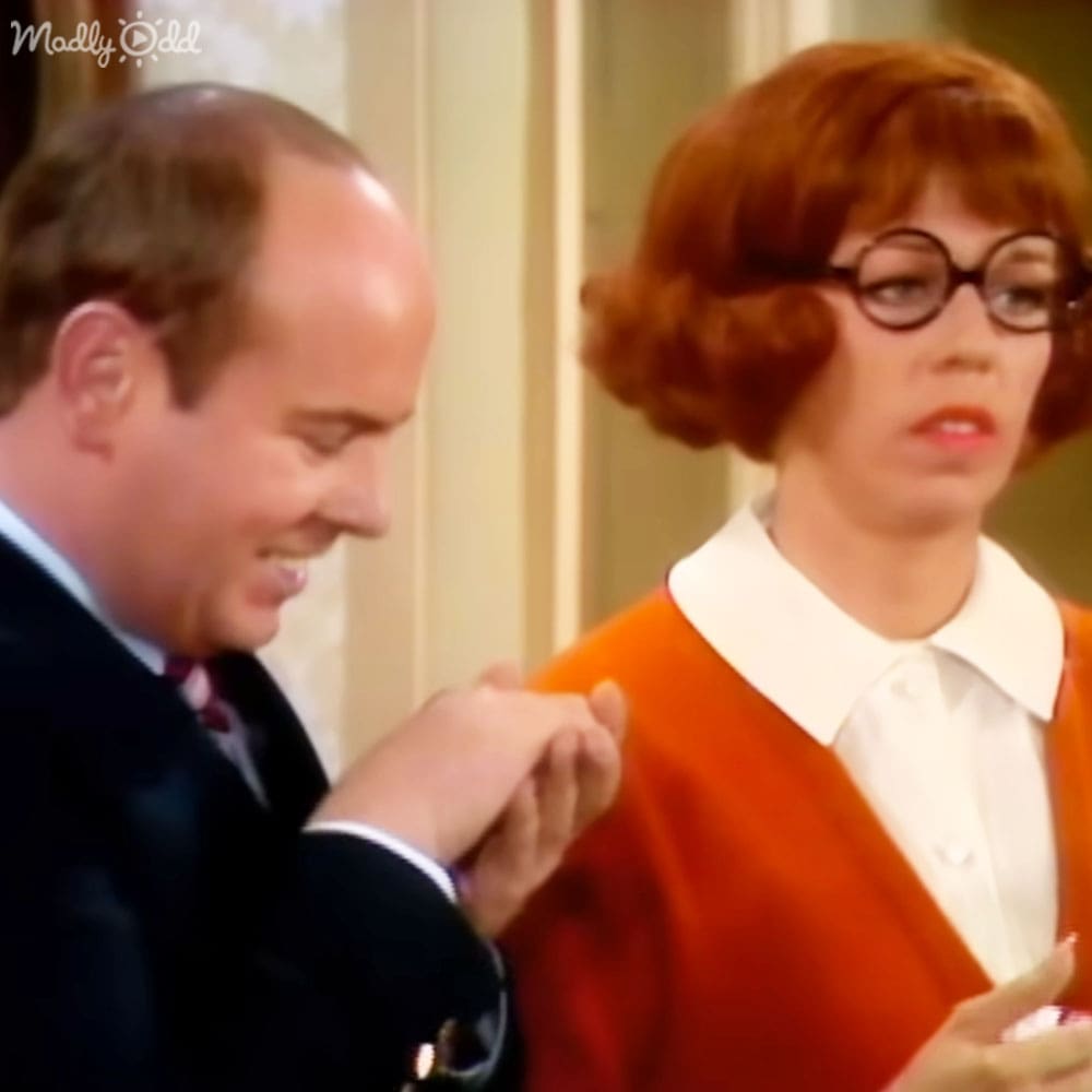 An adult male wearing a dark suit jacket with white trim on lapels, potentially from the 1960s or early 1970s, holding what appears to be a ring box. An adult female in an orange sweater and white collared shirt stands nearby.