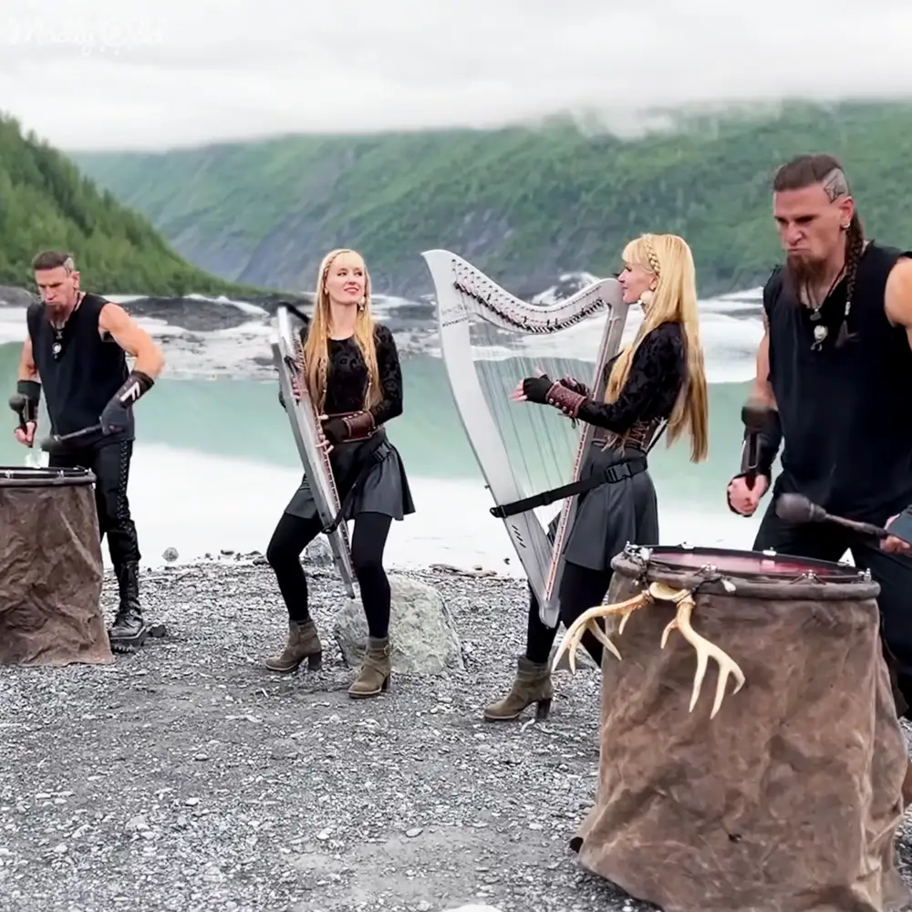Outdoor performance near a body of water with hills in spring or summer; group wearing coordinated dark-patterned attire performing music with harps and drums.