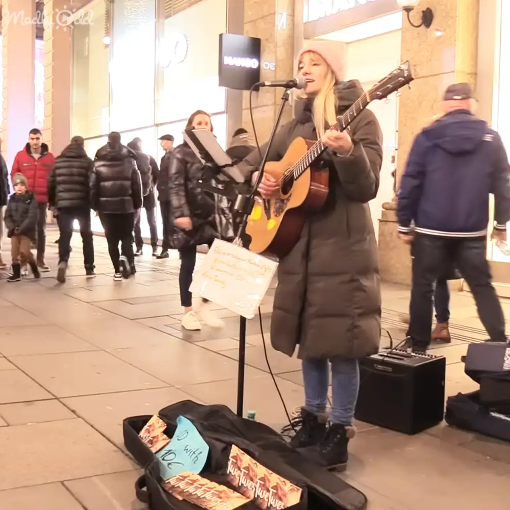 A young adult female street performer wearing a long dark puffer jacket, blue jeans, black ankle boots with fur trim, and a pinkish beanie hat plays guitar in an urban pedestrian area with storefronts. Pedestrians walk by, and a handwritten sign reads '10€' next to her open guitar case.