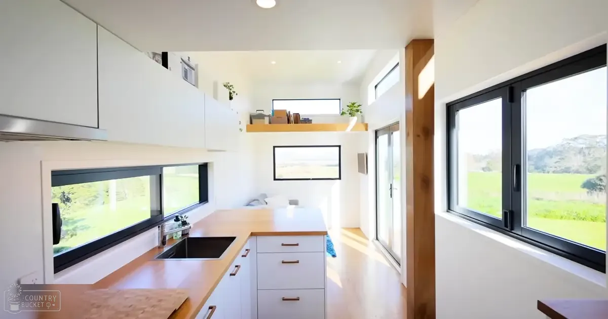 A modern kitchen interior with white cabinets, wooden countertops, a sink area, large windows offering views of lush greenery outside, and a framed picture on the wall in daylight within a rural house or cabin setting.