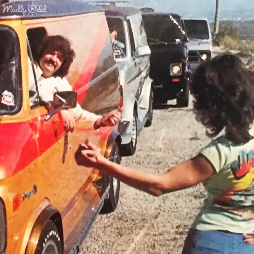 A young adult male and female stand beside parked vehicles in a dry, open area under clear daylight.