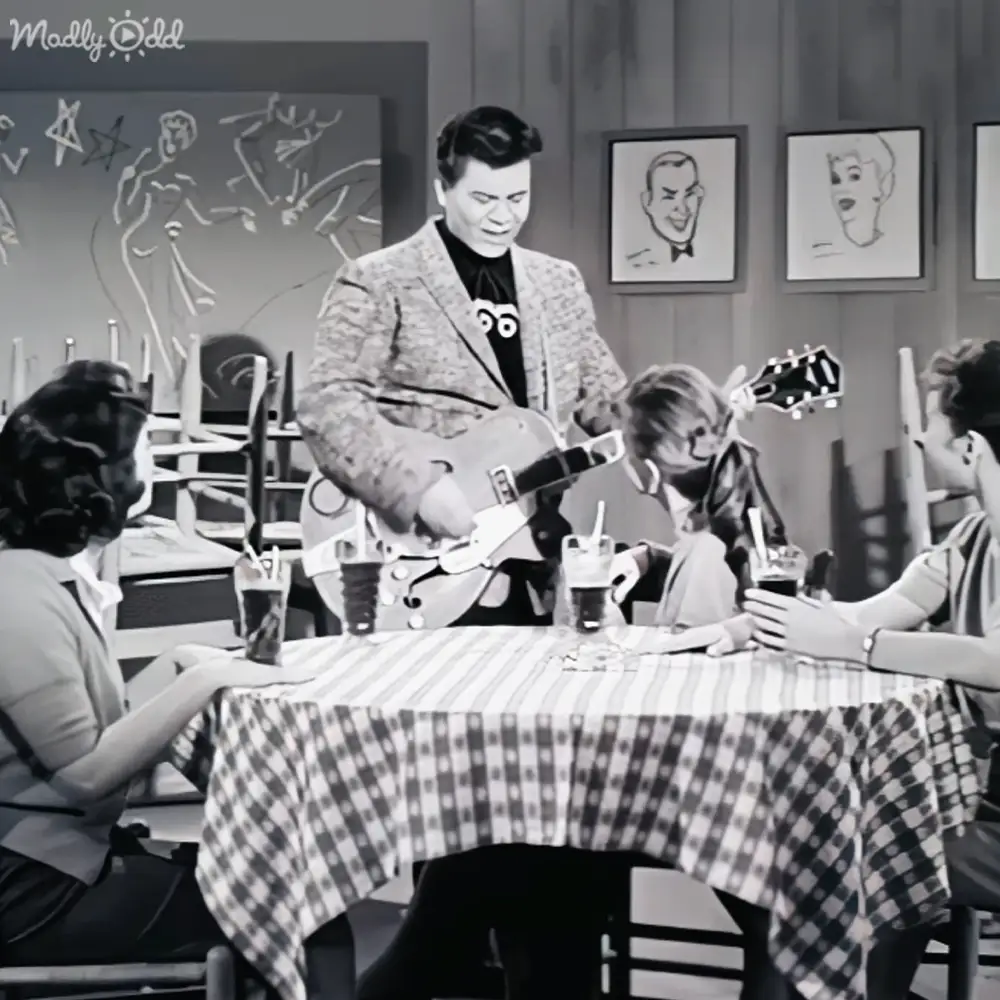 Adult male singer playing guitar in a diner or restaurant setting with checkered tablecloth, glasses of soda on the table, and stylized wall art in black-and-white.