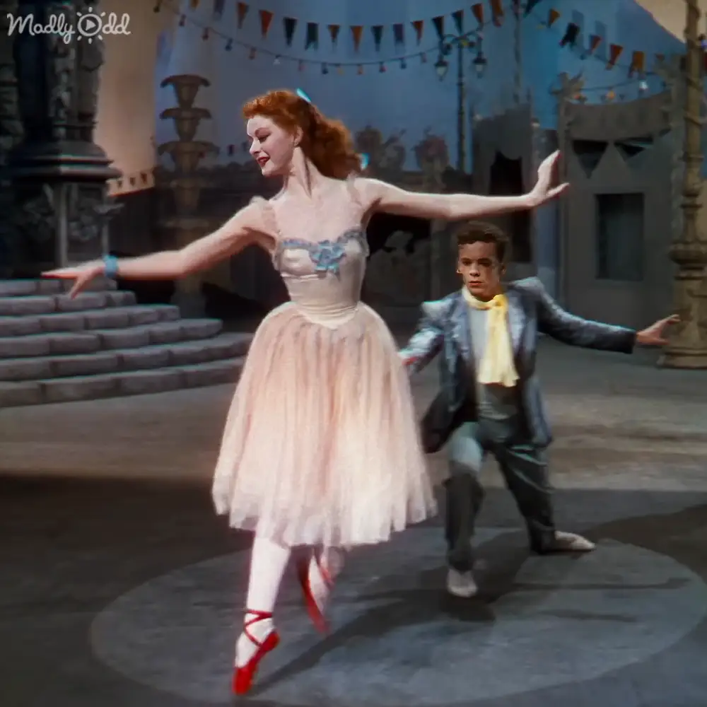 A ballet performance scene with a female dancer in a light pink tutu and male spectator in a dark suit on an indoor stage adorned with flags, columns, and steps.