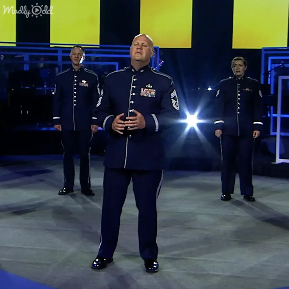Middle-aged man in a dark blue military-style uniform stands at center stage with two other individuals, flanked by microphones and professional stage lighting.