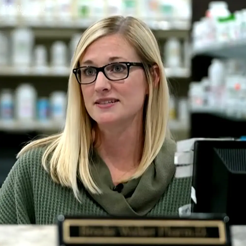 A middle-aged adult female wearing a green textured sweater with relaxed fit and V-neck, holding black-rimmed glasses, stands in an indoor pharmacy setting. Shelves stocked with medication or pharmacy supplies are visible.