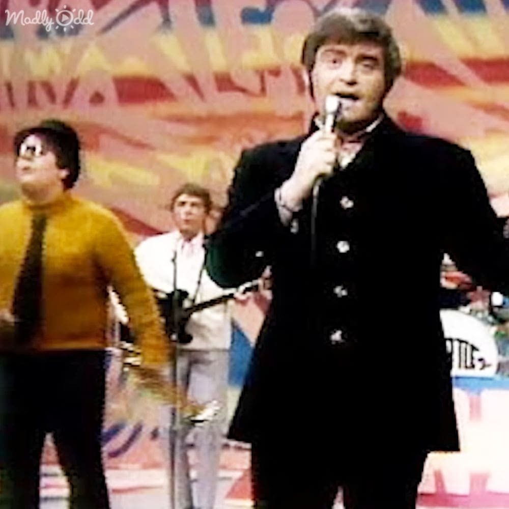A performer in a dark double-breasted coat sings into a microphone at an indoor concert venue, with other performers in formal attire behind him.