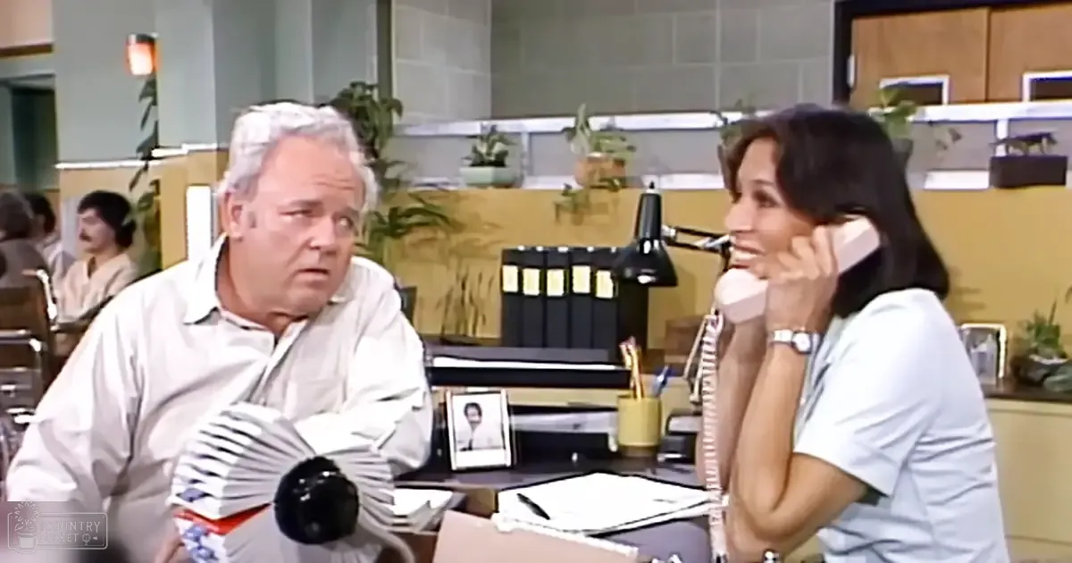 An adult male wearing a light-colored, long-sleeved 1970s-style shirt stands in an office setting with cubicles, desks, and plants around him.